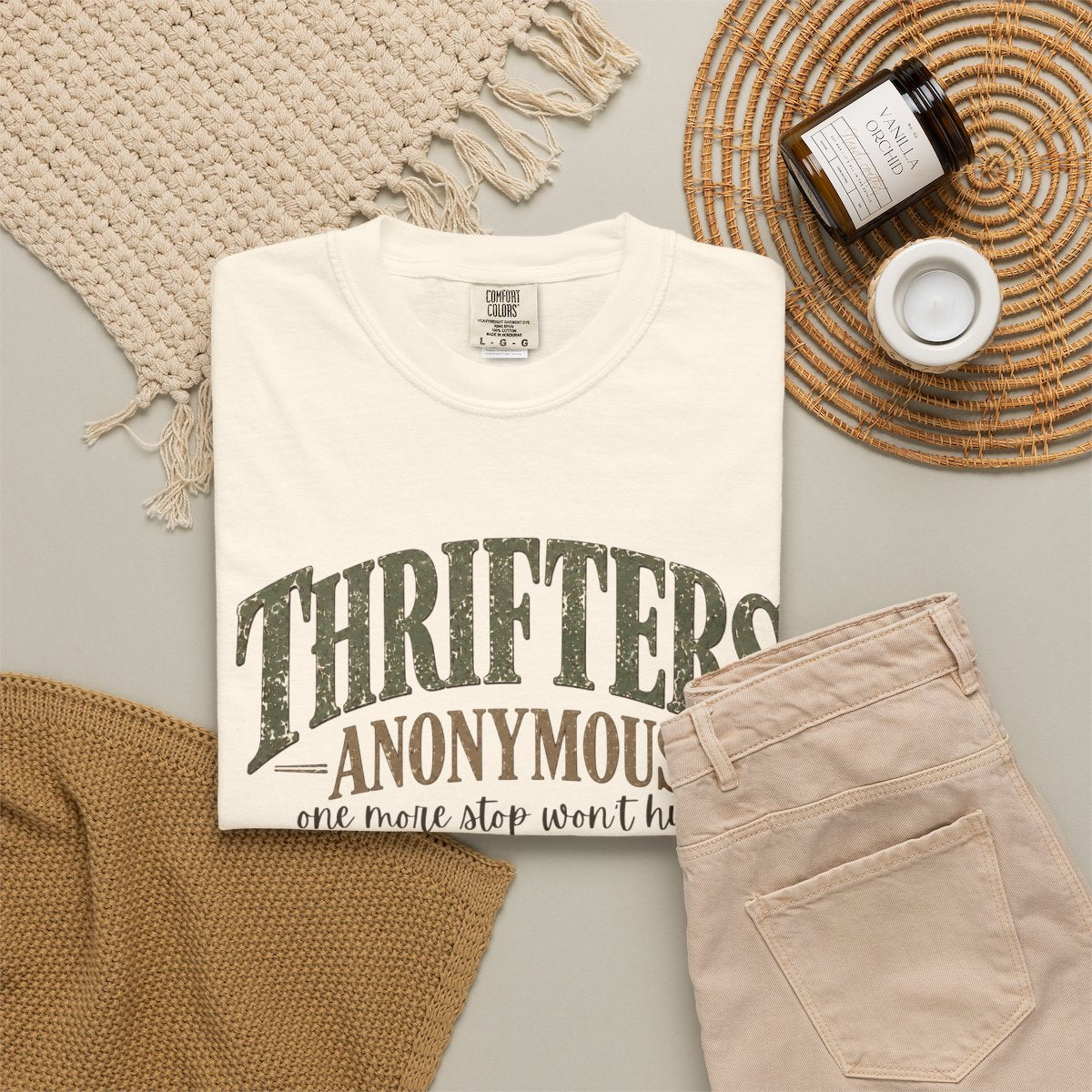 A folded cream-colored t-shirt with a distressed green graphic reading 'THRIFTER ANONYMOUS' and a quote underneath, styled in a flat lay with beige pants, a woven placemat, a candle jar, and a knit blanket on a light gray surface.