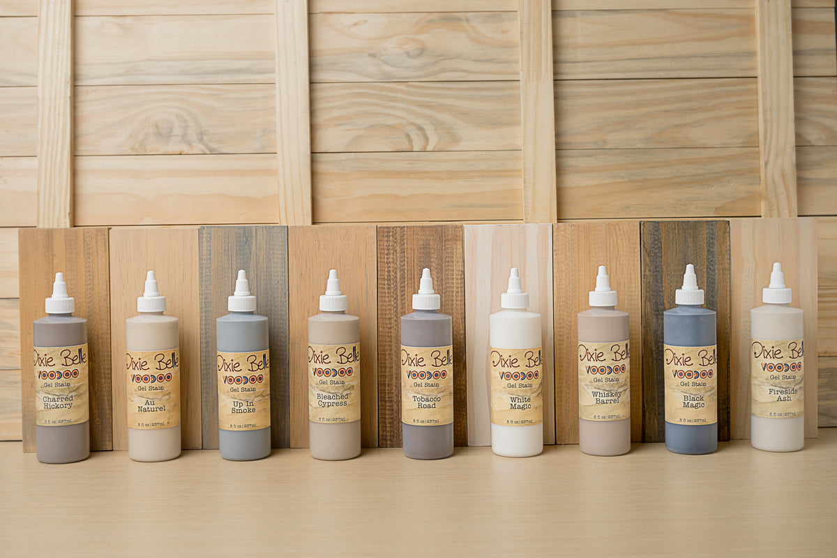 Nine bottles of VooDoo Gel Stain wood finish in various wood tones, arranged in a row against a light wood panel background.