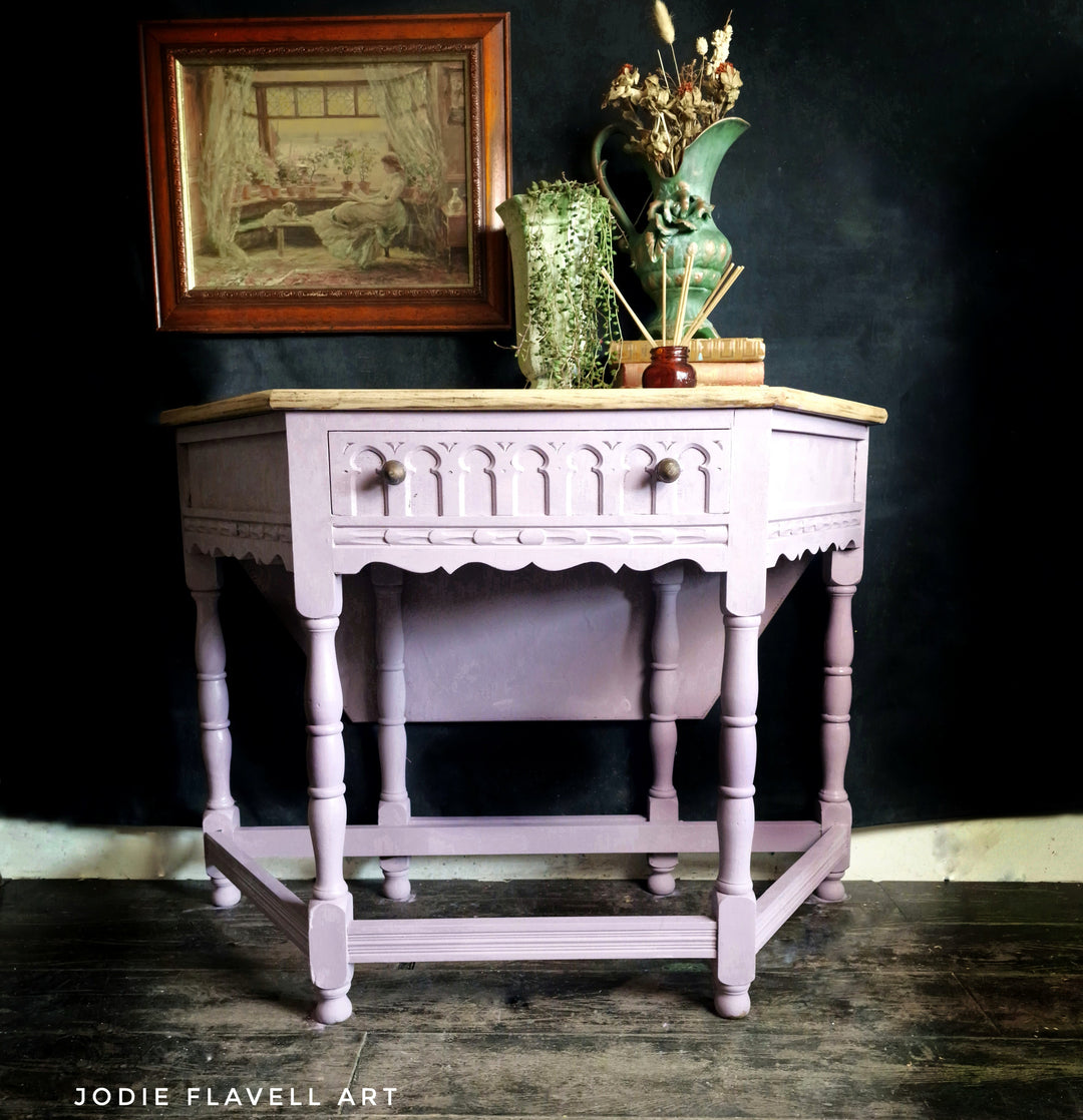 A vintage-style wooden writing desk painted in a dusty lavender-pink finish, featuring a light wood top, a single drawer with decorative knobs, and turned legs. It is styled with a green ceramic vase and books on top, set against a dark background.