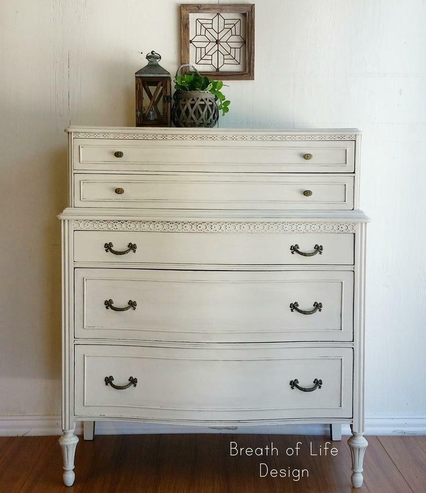 A distressed white wooden chest of drawers with five drawers, featuring dark metal handles and turned legs, set against a white wall with decorative items on top.