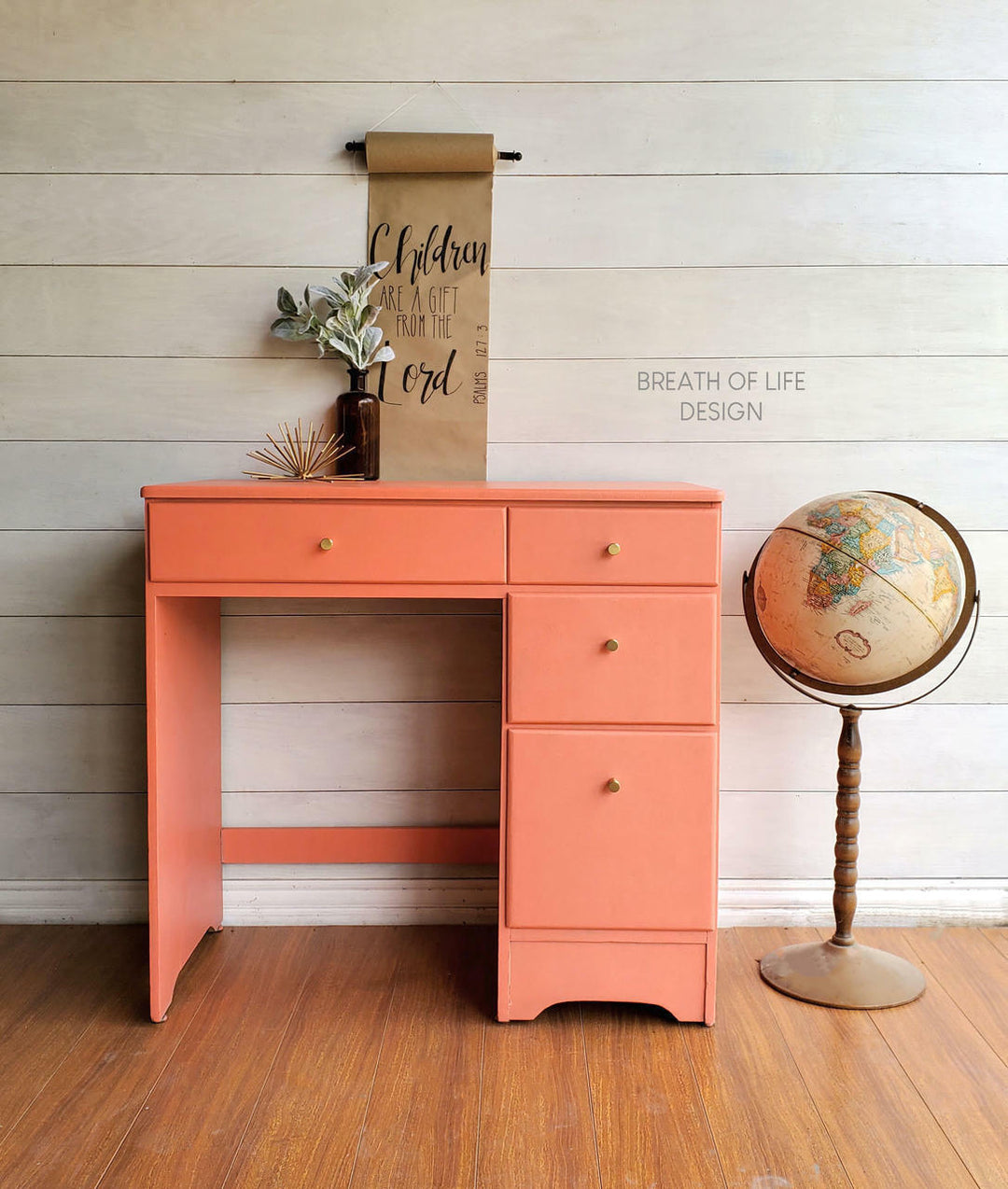 A coral pink wooden writing desk with three drawers and small brass knobs, styled in a room with a white shiplap wall, wood floor, a hanging scroll, and a vintage globe.