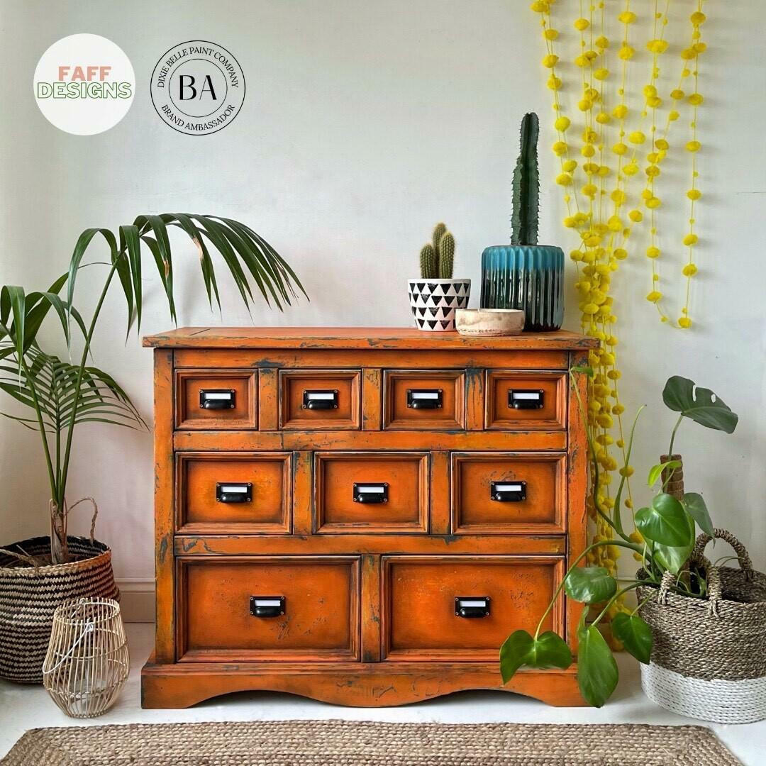 An orange-brown wooden 9-drawer chest of drawers styled in a home interior setting with potted plants, a cactus, and decorative hanging garlands.
