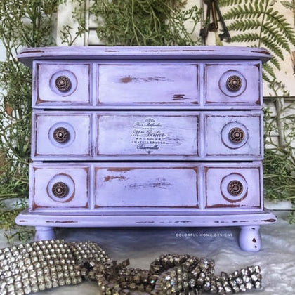 A distressed lavender purple three-drawer chest of drawers with round metal knobs, styled with a beaded garland in front and green foliage in the background.