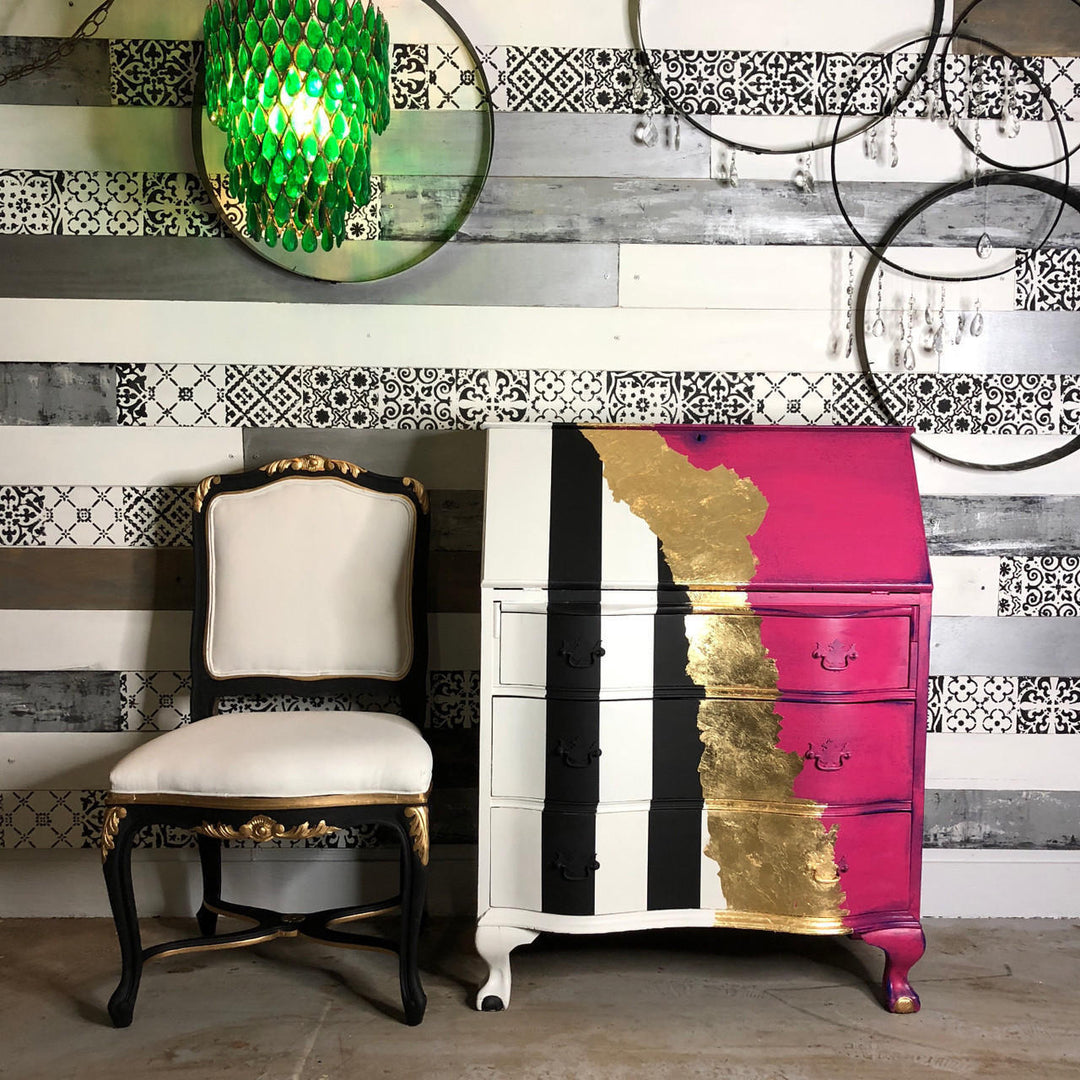 A painted three-drawer chest of drawers with a bold vertical color-blocked design in white, black, and bright pink, accented with gold leaf on the right side. It is styled in a room setting next to a matching black and cream chair, with a patterned wall and hanging glass ornaments in the background.