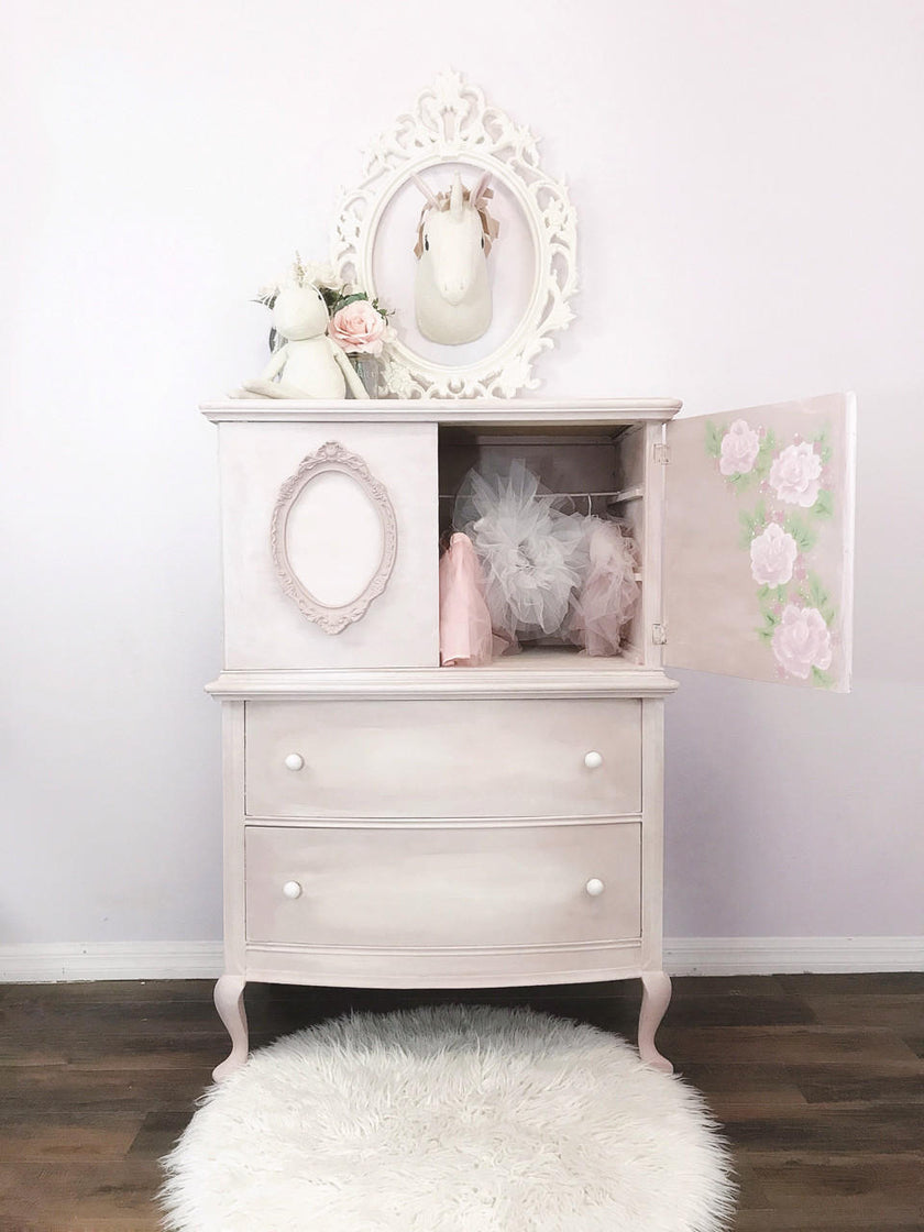 A light pink distressed wooden hutch with two drawers and an open door showing pink tulle skirts inside. The hutch is decorated with a white unicorn figurine, a floral mirror frame, and a unicorn head plaque, set against a white wall and dark wood floor with a white rug.