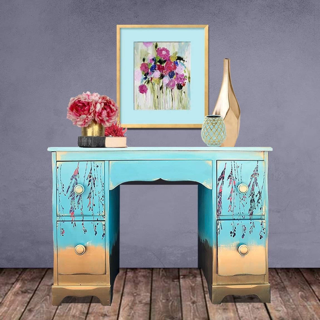 A painted writing desk with a turquoise and gold finish, decorated with floral motifs on the drawer fronts, styled with a framed painting, a vase, and a small pot against a textured gray wall and wooden floor.