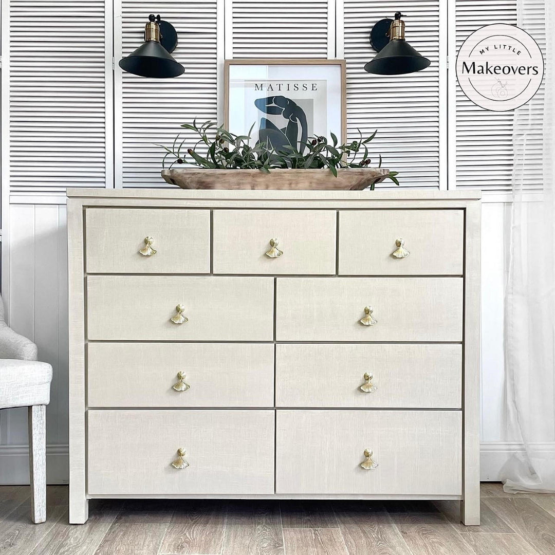 An off-white linen-textured 7-drawer dresser with gold-toned knobs, styled in a room setting with a decorative tray of greenery on top, black wall sconces, and a white shuttered wall background.