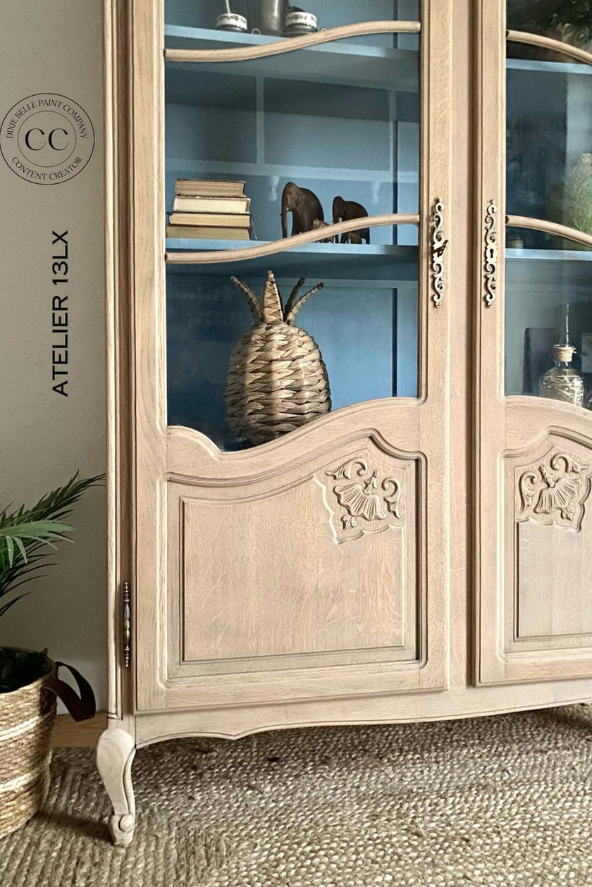 A tall, light beige carved wooden display cabinet with glass doors, shown in a styled room setting with a woven rug, a potted plant, and decorative items inside.