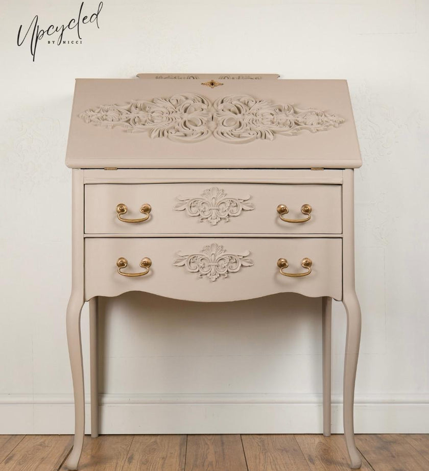 A small writing desk with two drawers, ornate floral carvings, and curved legs, shown against a plain wall on a wood floor. The desk is painted in a warm light beige/taupe tone with gold-toned drawer pulls.