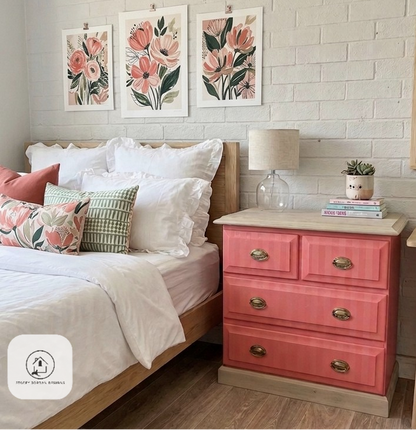 A bedroom scene showing a coral pink three-drawer nightstand with light wood trim next to a bed with white bedding and floral accent pillows. A glass table lamp sits on top of the nightstand, which is set against a white painted brick wall with three matching floral wall art prints.