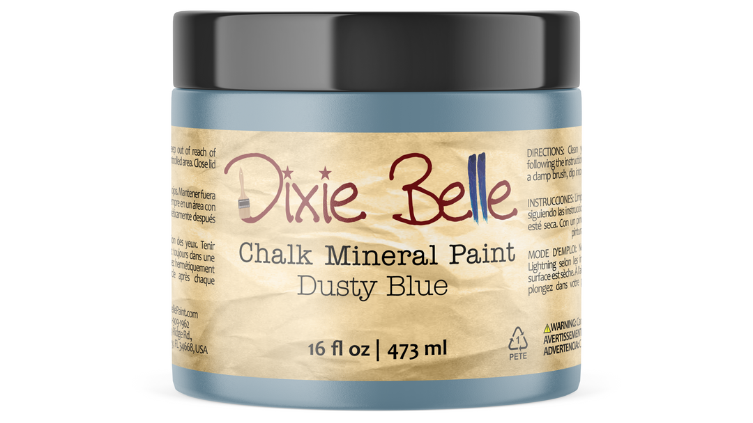 A jar of Dixie Belle Chalk Mineral Paint in the color 'Dusty Blue', featuring a light blue-gray container, a black lid, and a beige paper label with red and brown lettering.