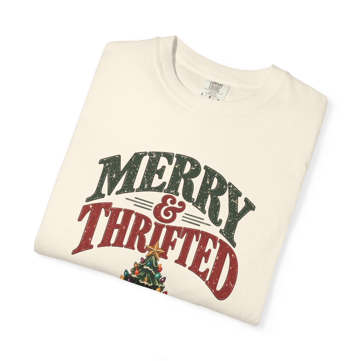 A folded cream-colored t-shirt with a distressed graphic that reads 'MERRY & THRIFTED' in dark green and red lettering, with a small Christmas tree illustration below, shown on a plain white background.