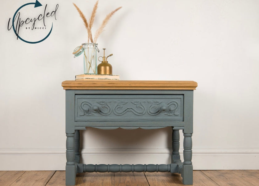 A painted wooden console table with a dusty blue-gray finish and a natural light wood top, styled with a glass vase, pampas grass, and a brass oil lamp against a white wall.