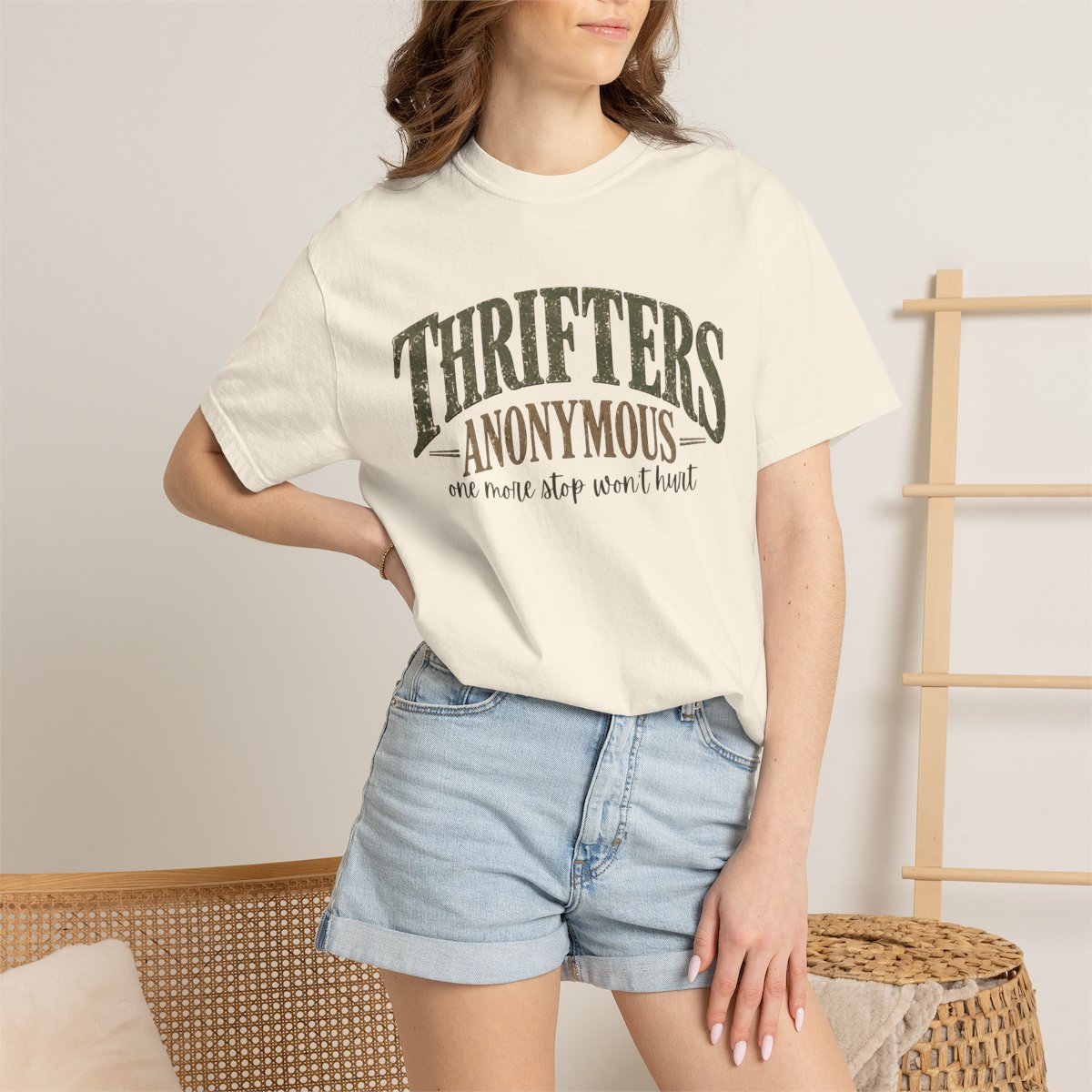 A woman wearing a cream-colored graphic t-shirt with 'THRIFTERS ANONYMOUS' printed in dark green and brown, paired with light-wash denim shorts in a cozy indoor setting.