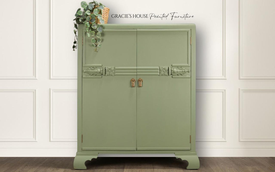 A sage green painted wooden cabinet with two doors, decorative paneling, and small gold-toned padlocks, shown against a white paneled wall with a wood floor. A hanging plant decorates the top left corner, and the text 'GRACIE'S HOUSE Painted Furniture' appears at the top.