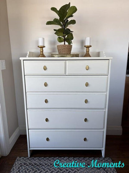 A white five-drawer dresser with round brass knobs, styled with a potted fiddle-leaf fig, candles, and gold candlesticks on top, set against a beige wall.