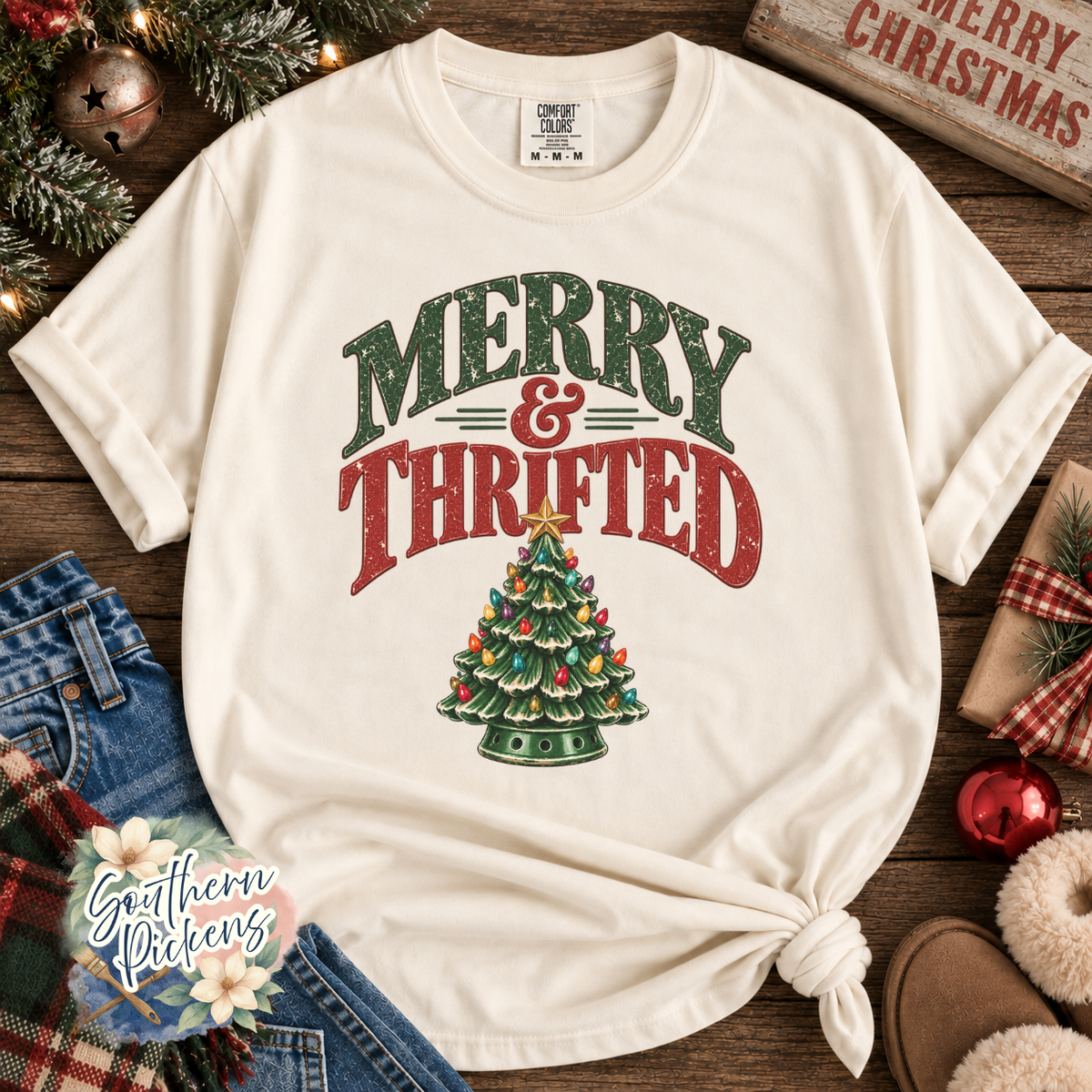 An off-white short-sleeve t-shirt with a distressed Christmas graphic reading 'MERRY & THRIFTED' above a decorated tree, styled in a flat-lay with jeans, holiday greenery, and a wooden sign.