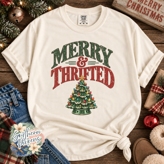An off-white short-sleeve t-shirt with a distressed Christmas graphic reading 'MERRY & THRIFTED' above a decorated tree, styled in a flat-lay with jeans, holiday greenery, and a wooden sign.