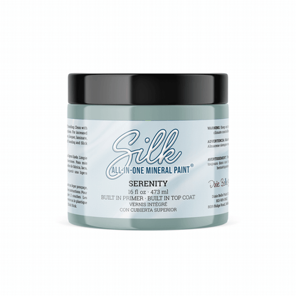 A 16oz. jar of Silk All-In-One Mineral Paint in the shade Serenity, featuring a light blue-gray body and a black lid, set against a plain white background.