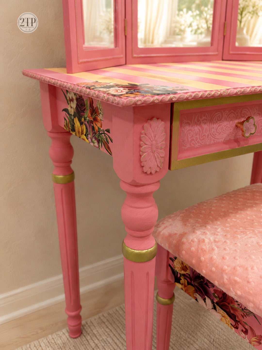 A pink vanity table with a floral and striped patterned top, matching pink pedestal legs with gold trim, and a coordinating pink upholstered bench in a styled interior setting.