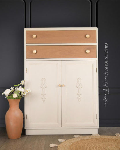 A tall painted wooden cabinet with two upper drawers, two lower doors, and small round knobs, shown in a styled interior setting with a vase of flowers and a woven rug against a dark paneled wall.