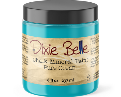 A jar of Dixie Belle Chalk Mineral Paint in the color 'Pure Ocean', featuring a bright turquoise blue paint inside, a black screw-on lid, and a beige paper label with red and blue lettering.