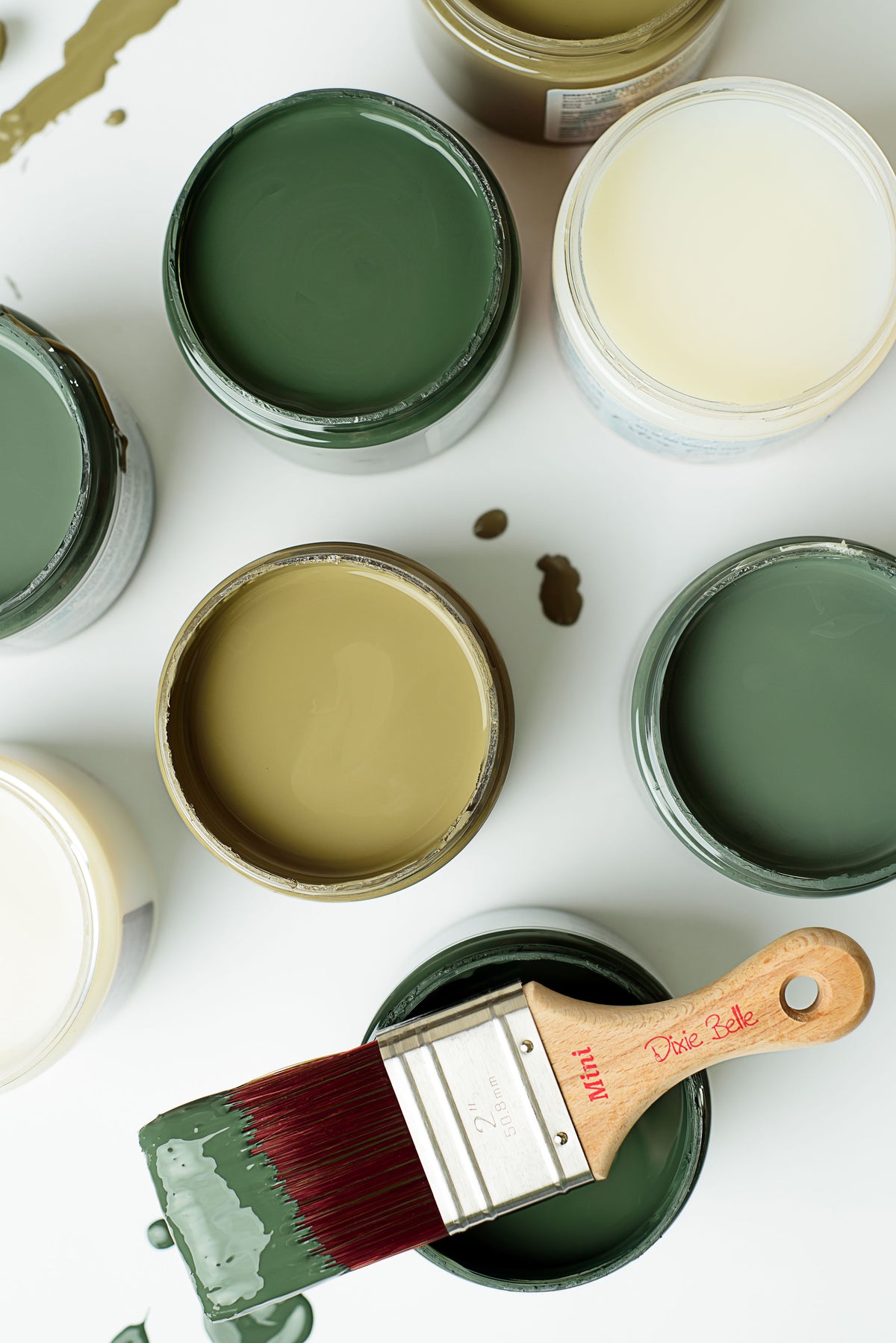 An overhead flat-lay of open paint cans in muted green, beige, and off-white shades, with a paintbrush resting in one can on a white surface.
