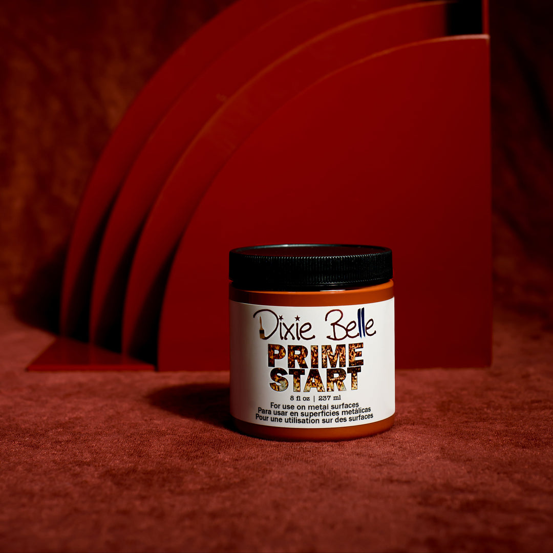 A jar of Dixie Belle Prime Start wood finish with a white label and black lid, set against a dark red textured background with matching red arches.
