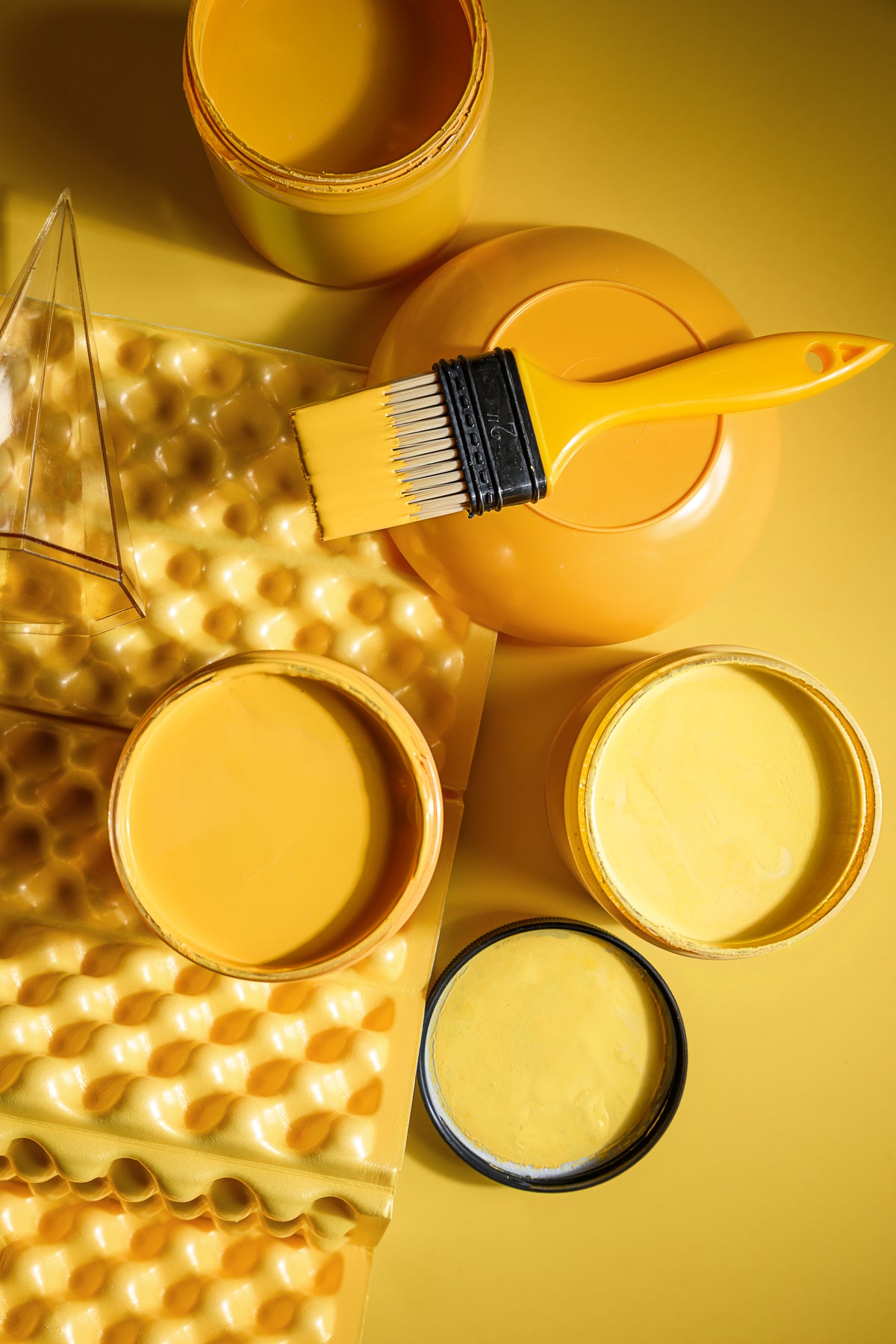 Yellow paint in multiple containers, a yellow paint brush, and a yellow textured sponge on a yellow background.
