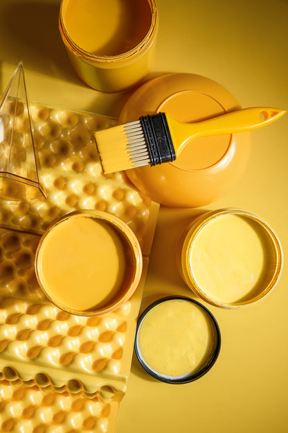 Yellow paint in multiple containers, a yellow paint brush, and a yellow textured sponge on a yellow background.
