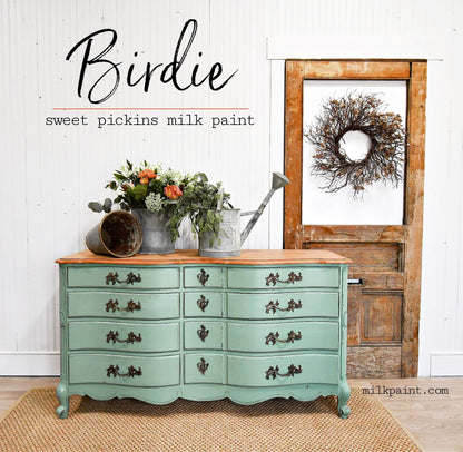 A painted vintage-style chest of drawers in a soft mint green with a natural wood top, styled in a rustic room setting with a wooden door, wreath, and metal watering can on a woven rug. Text on the image reads 'Birdie sweet pickins milk paint'.