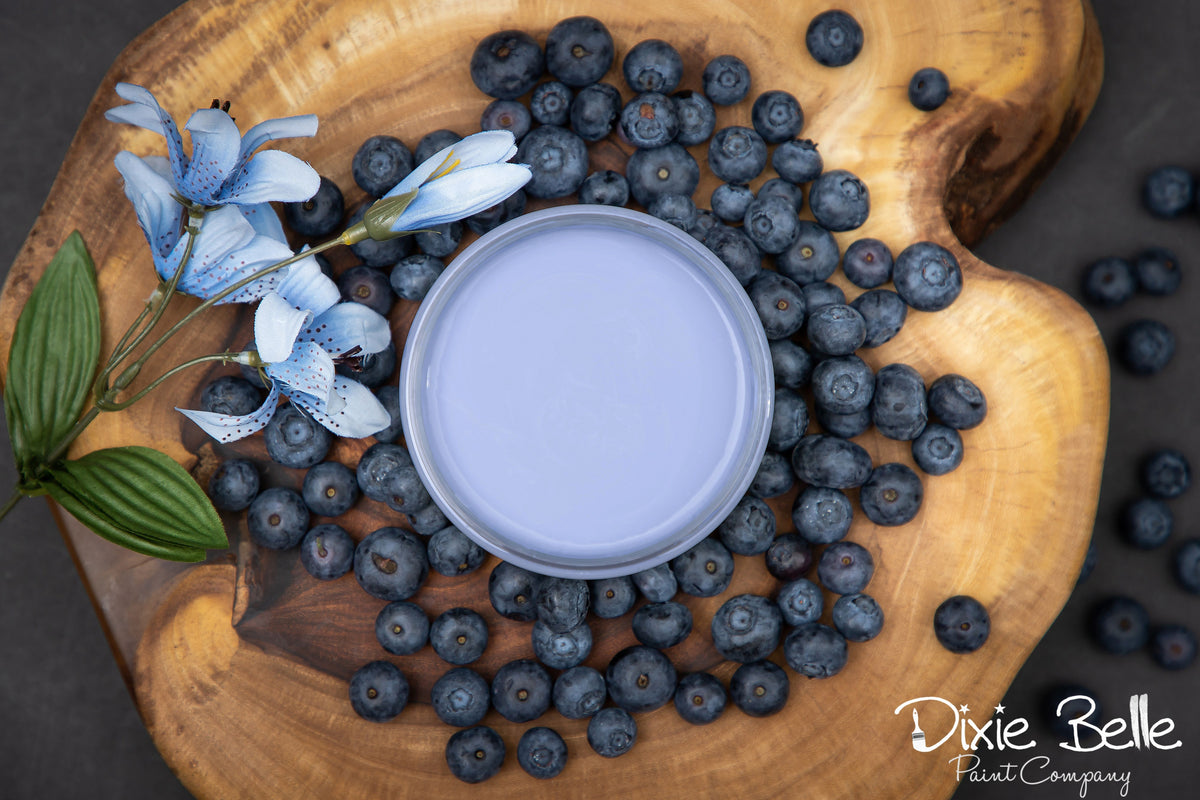 A round tin of light periwinkle blue paint sits on a rustic wooden board surrounded by fresh blueberries and blue flowers. The Dixie Belle Paint Company logo is visible in the bottom right corner.