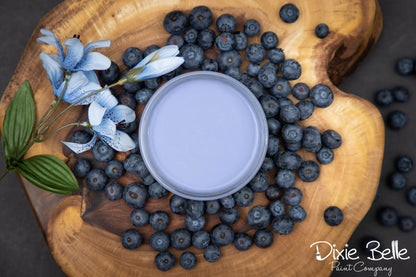 A round tin of light periwinkle blue paint sits on a rustic wooden board surrounded by fresh blueberries and blue flowers. The Dixie Belle Paint Company logo is visible in the bottom right corner.