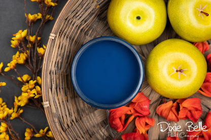 Blue paint jar in a wicker basket with green apples and red flowers on a dark surface