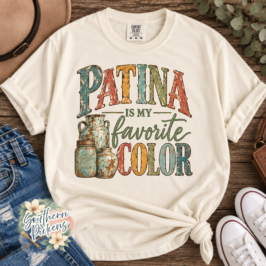 An off-white graphic t-shirt styled in a flat-lay arrangement on a rustic wooden background, featuring the text 'PATINA IS MY favorite COLOR' in a distressed, multi-colored font alongside vintage mason jar illustrations. The shirt is knotted at the bottom hem and surrounded by jeans, white sneakers, a hat, and eucalyptus leaves.