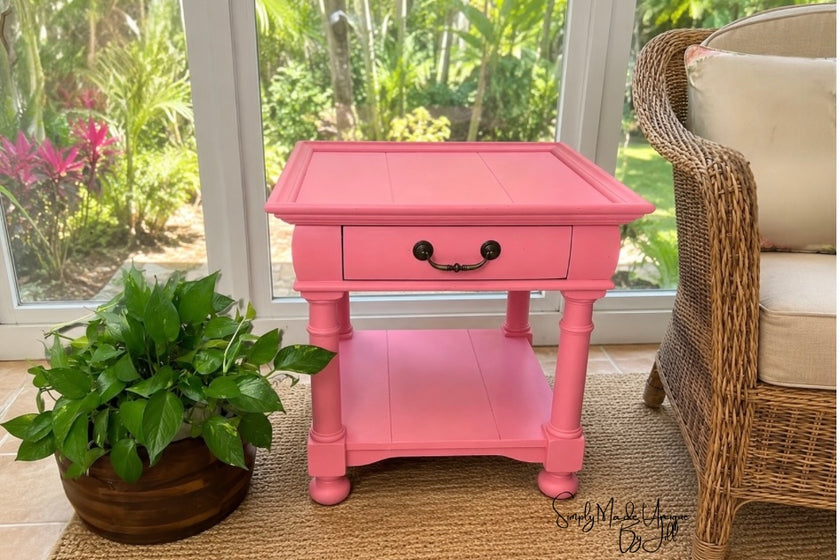 A bright pink wooden end table with a single drawer and lower shelf, styled in a sunroom next to a wicker chair and a potted plant.