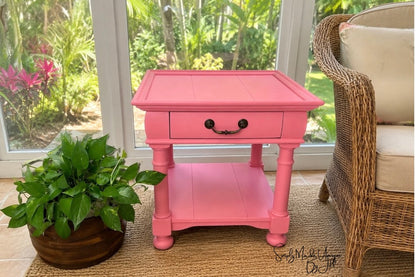 A bright pink wooden end table with a single drawer and lower shelf, styled in a sunroom next to a wicker chair and a potted plant.