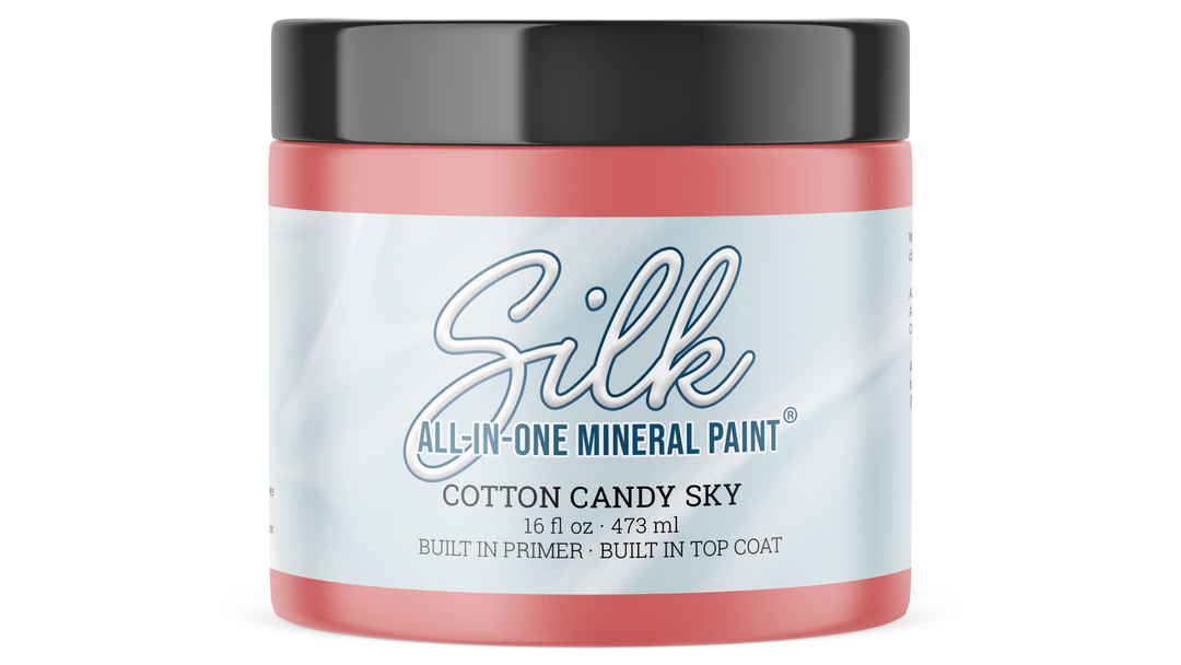 A jar of Silk All-In-One Mineral Paint in the color Cotton Candy Sky, featuring a light blue and white label with blue text, a coral pink base, and a black lid.