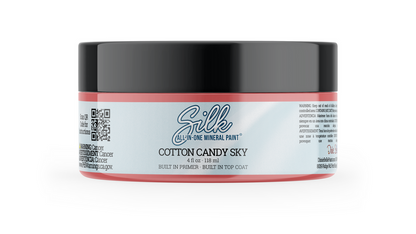 A jar of Silk All-In-One Mineral Paint in the color 'Cotton Candy Sky', featuring a black lid, a coral pink rim, and a light blue label with white text.
