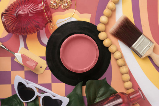 A styled flat-lay featuring a container of Cotton Candy Sky Silk Paint, surrounded by colorful props including a red ribbed glass container, wooden beads, a paintbrush, heart-shaped sunglasses, green leaves, and a patterned orange and purple background.