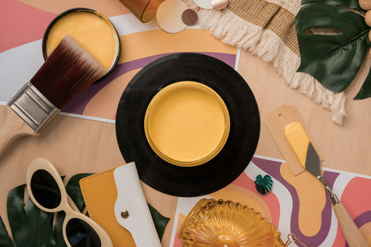 A styled flat-lay featuring a round black tray or plate filled with bright yellow Golden Hour Silk Paint, surrounded by decorative props including a paintbrush, sunglasses, a small fan, a notebook, and tropical leaves on a pastel abstract background.