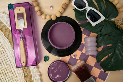 A styled flat-lay featuring Heaven Sent Silk Paint with a metal palette tool in a purple box, a round tin of purple pigment, an open jar, a brush, sunglasses, wooden beads, green leaves, and a checkered mat on a light wood surface.