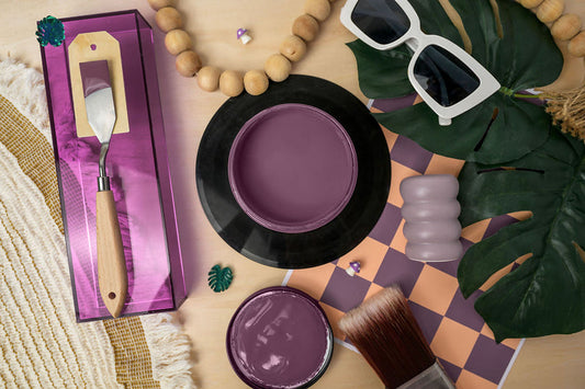 A styled flat-lay featuring Heaven Sent Silk Paint with a metal palette tool in a purple box, a round tin of purple pigment, an open jar, a brush, sunglasses, wooden beads, green leaves, and a checkered mat on a light wood surface.