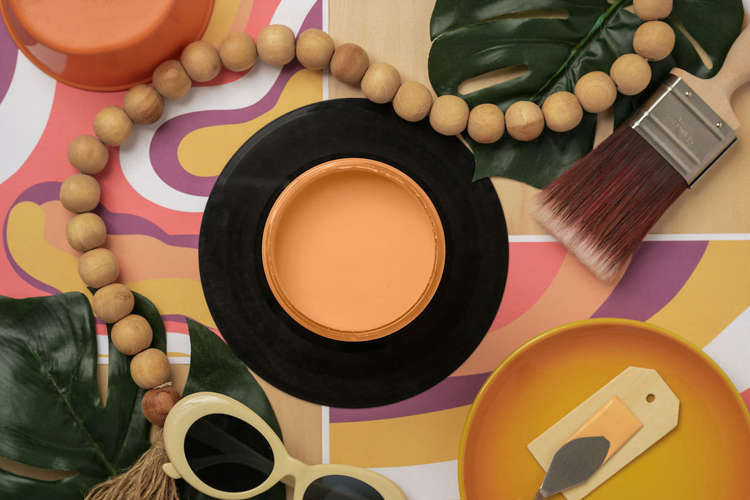 A styled flat-lay featuring Sunset Horizon Silk Paint and a black casing, surrounded by tropical leaves, wooden beads, a paintbrush, sunglasses, and a yellow palette on a colorful abstract patterned background.