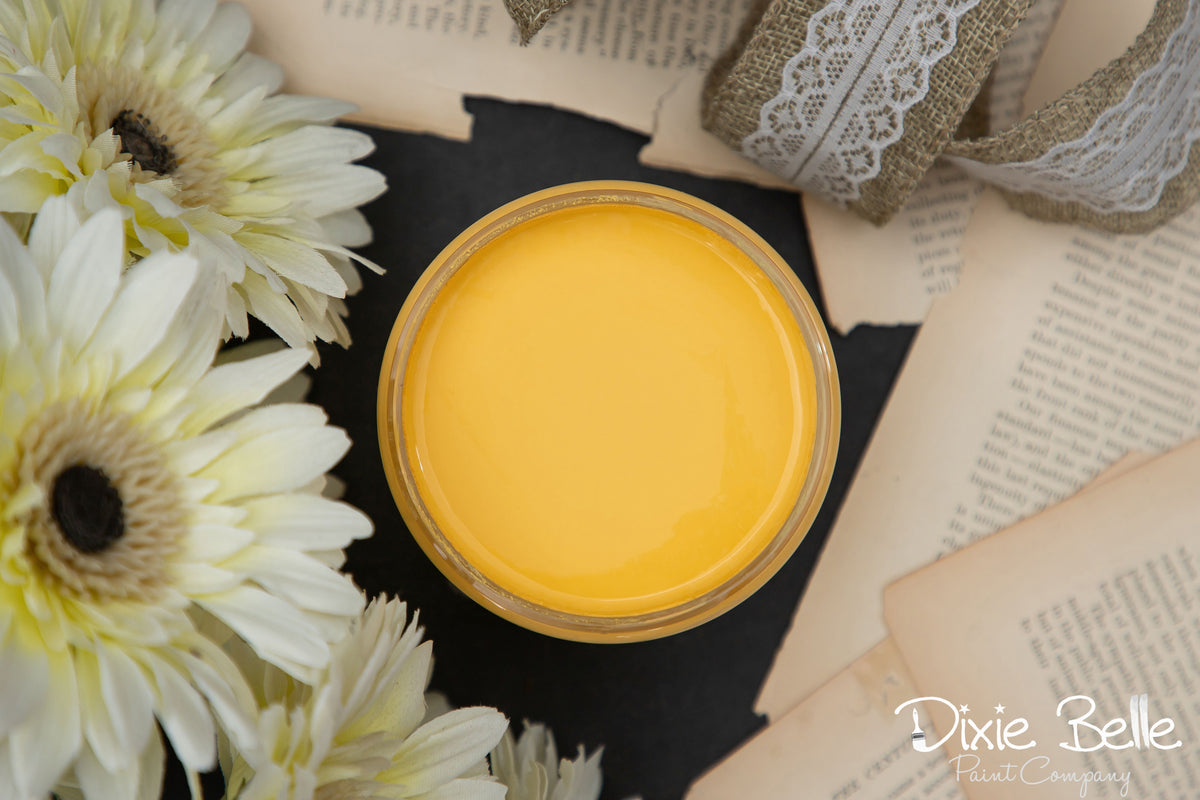 Jar of yellow paint with flowers and lace on a black surface, featuring the brand 'Dixie Belle'.
