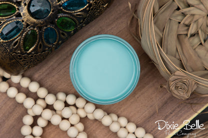 A round open jar of light aqua blue paint, styled on a wooden surface with decorative beads, a woven palm leaf piece, and an ornate jeweled object. The Dixie Belle Paint Company logo is visible in the bottom right corner.