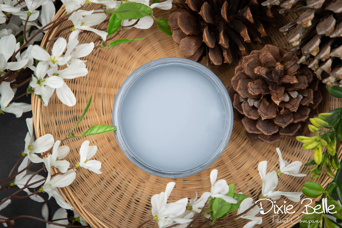 Round container of dusty blue painy with a lid on a woven mat surrounded by white flowers and pine cones, featuring the brand 'Dixie Belle'.