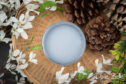 Round container of dusty blue painy with a lid on a woven mat surrounded by white flowers and pine cones, featuring the brand 'Dixie Belle'.