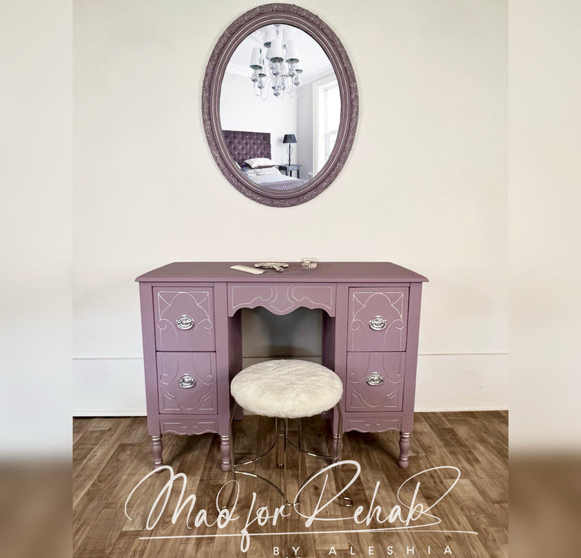 A lavender purple vanity desk with ornate silver drawer pulls, an open center compartment, and a round tufted cream stool underneath. Above it is an oval mirror with a matching purple frame, set against a beige wall on a wood floor.