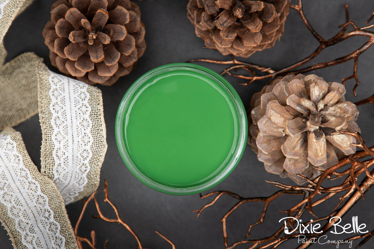 Green paint jar surrounded by pine cones and branches on a dark surface with 'Dixie Belle' branding.